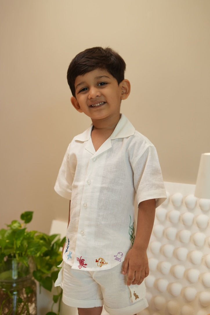 Child wearing a white embroidered outfit standing against a beige wall with a plant in the background.