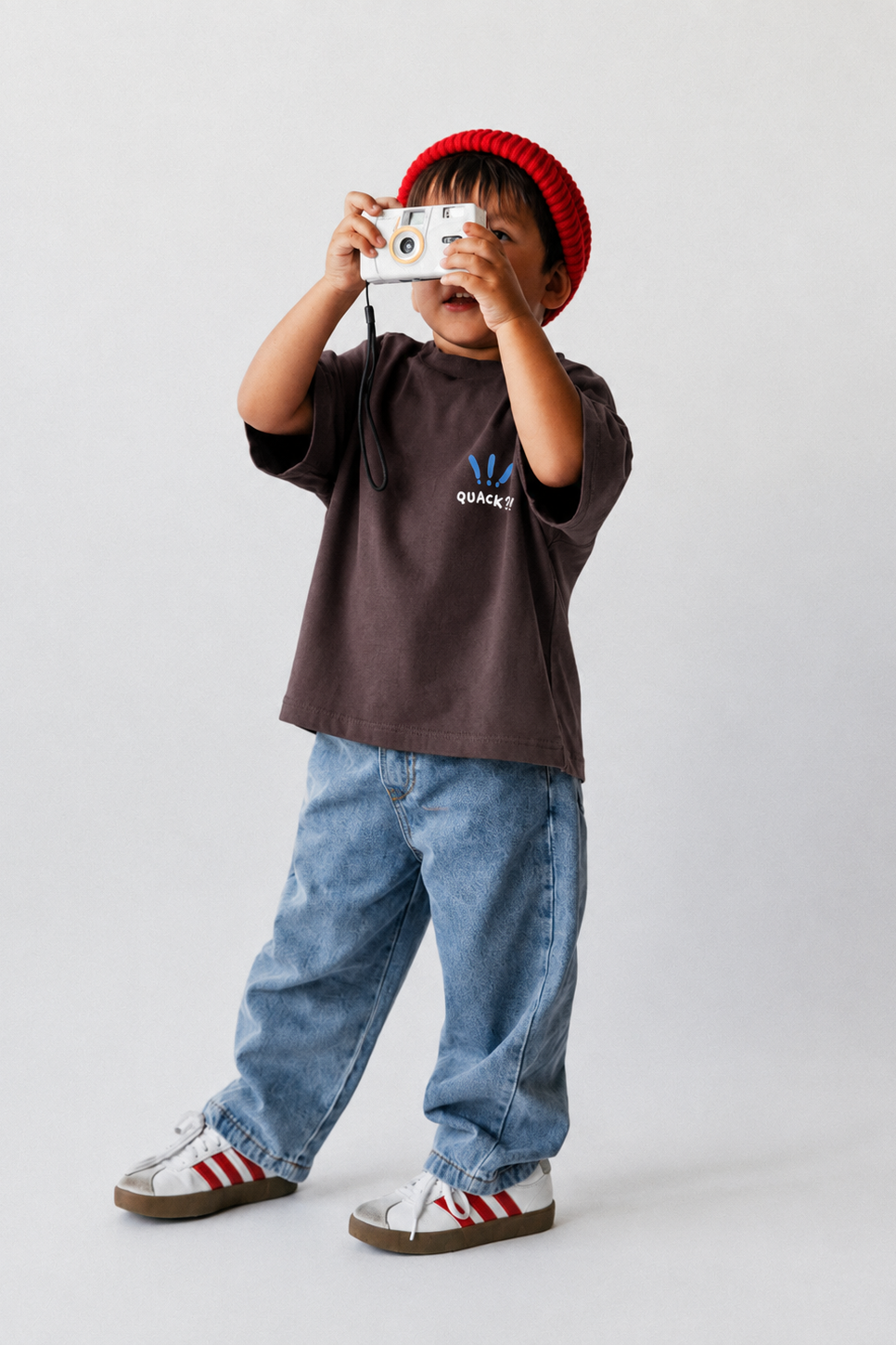 Child wearing a brown t-shirt with a logo, blue jeans, and a red knit hat holding a camera.