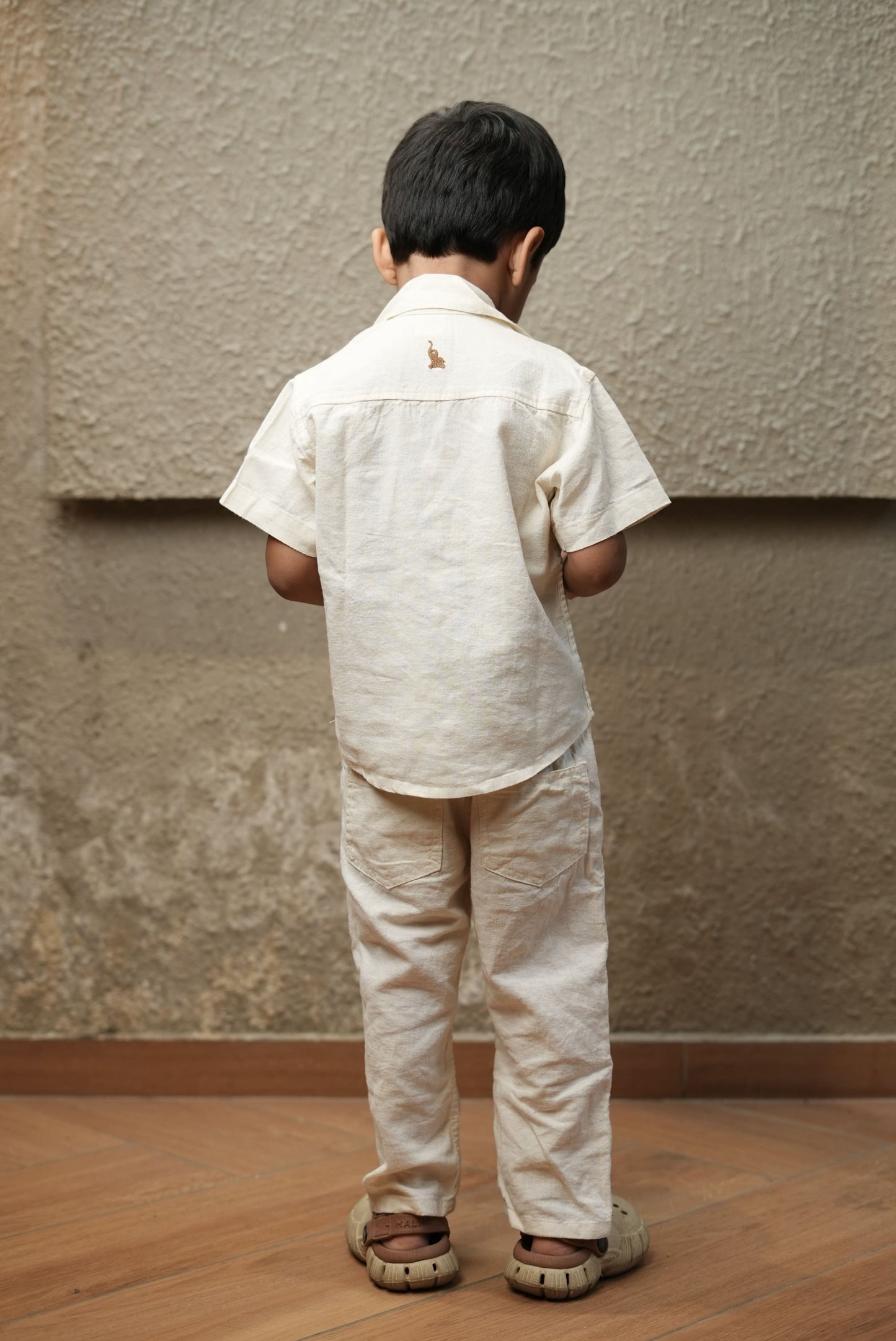 Child wearing a beige shirt and pants standing against a textured wall.