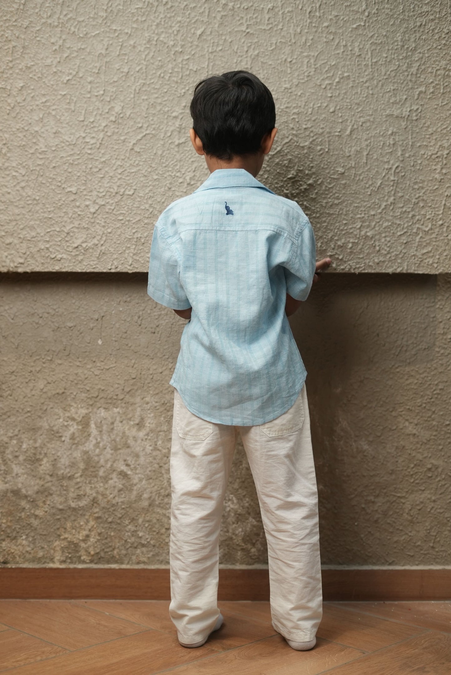 Child in a light blue shirt and white pants standing against a textured wall.