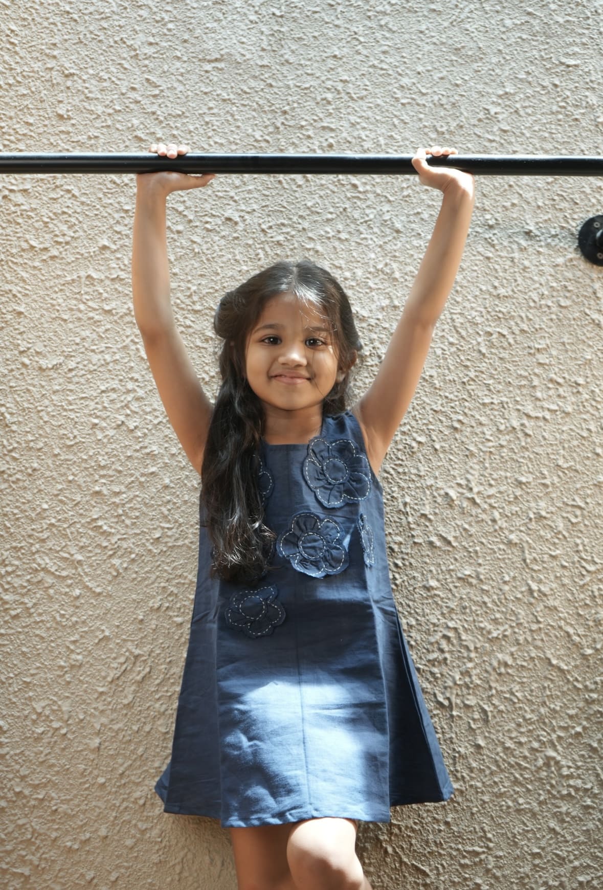 Young girl in a blue dress hanging from a horizontal bar against a beige wall.