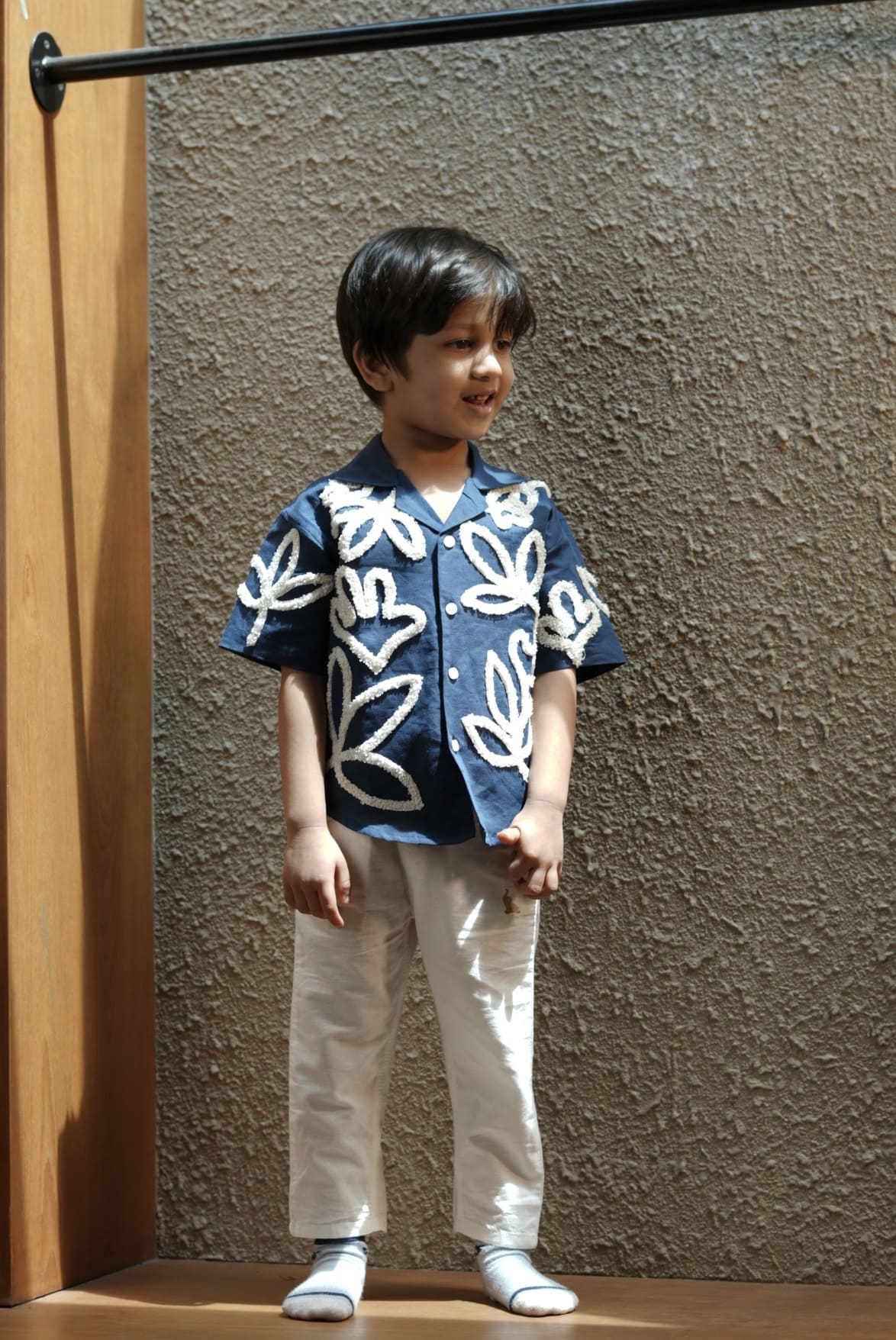 Child wearing a blue floral shirt and white pants standing against a textured wall.