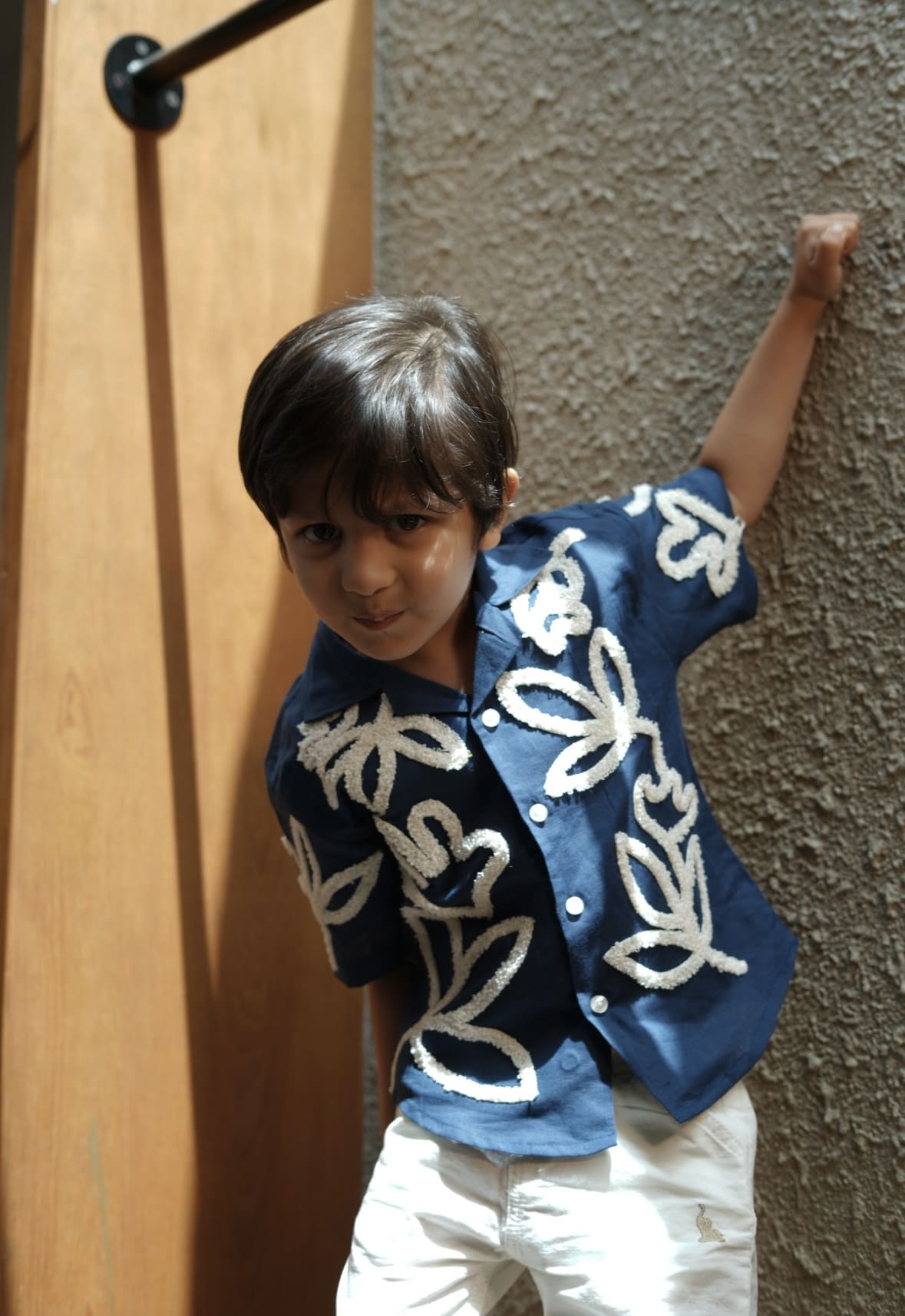 Child wearing a blue embroidered shirt against a textured wall.
