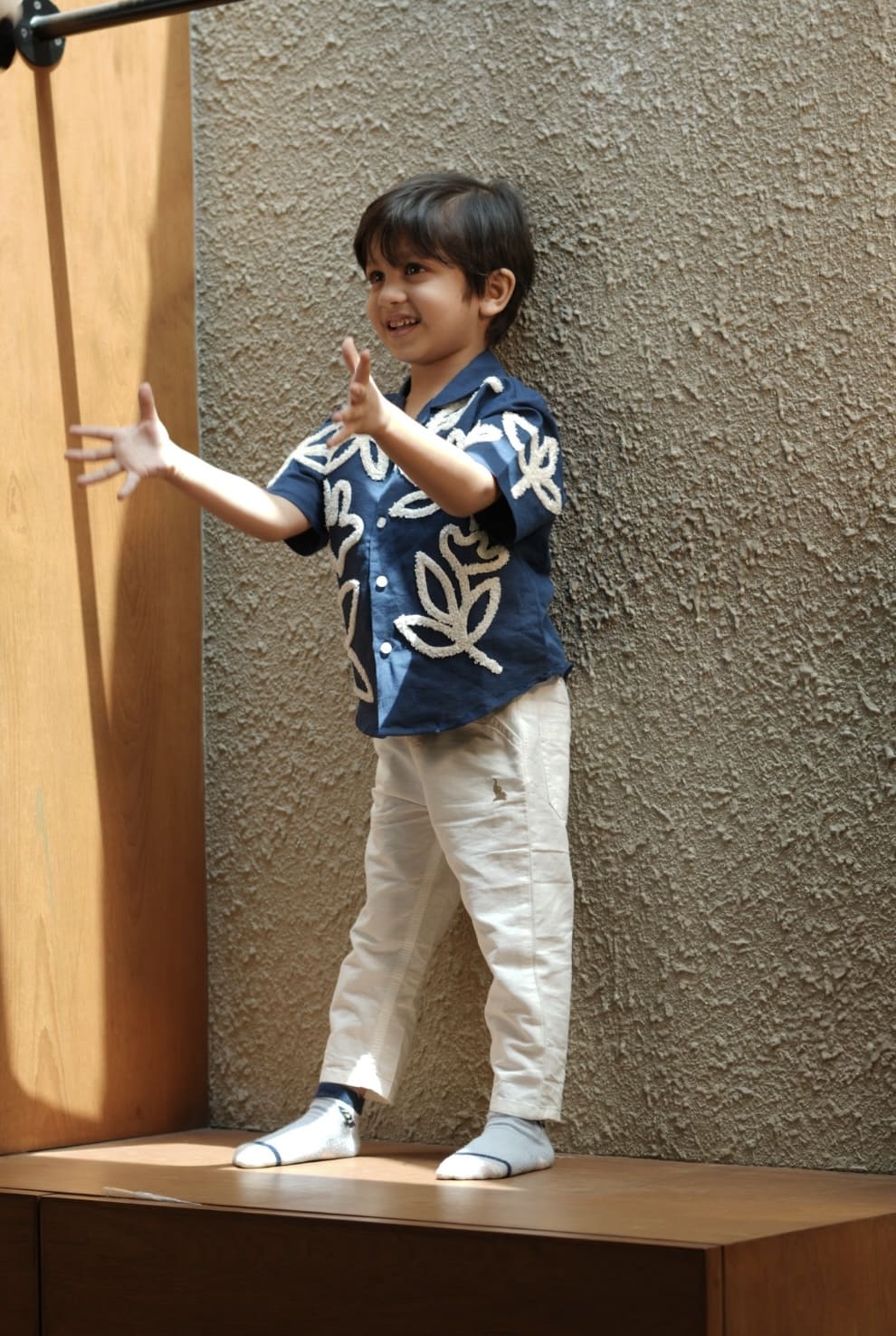 Child wearing a blue shirt with white patterns and white pants standing on a wooden platform against a textured wall.