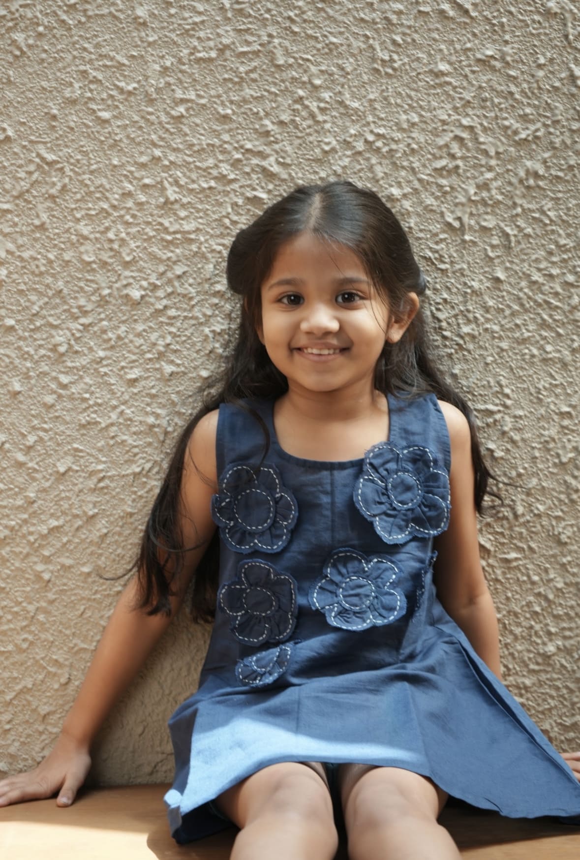 Young girl wearing a blue dress with floral patterns against a beige wall.
