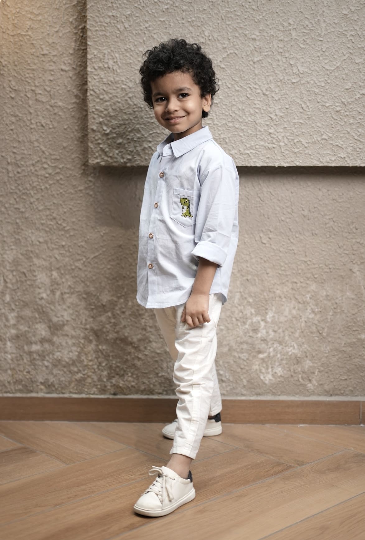 Child wearing a light-colored shirt and white pants standing against a textured wall.