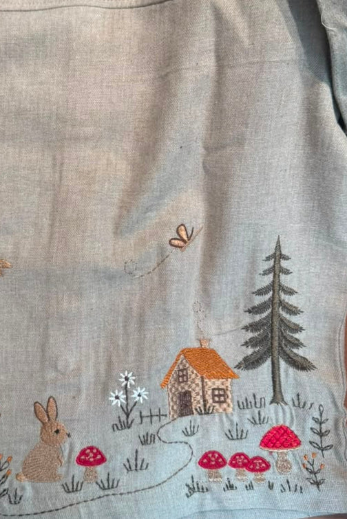 A light beige linen shirt with colorful embroidered woodland motifs, including a fox, rabbit, house, trees, and mushrooms, laid flat on a wooden surface.