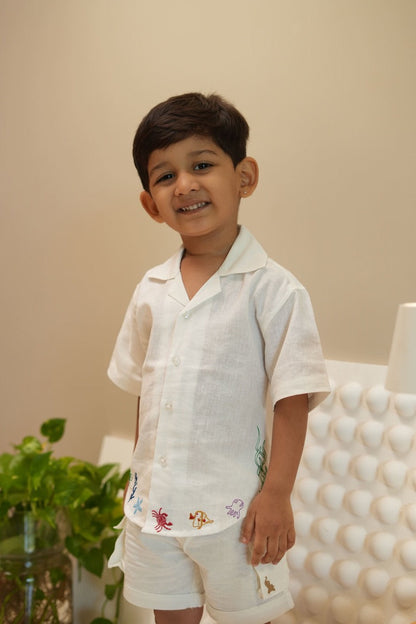Child wearing a white embroidered outfit standing against a beige wall with a plant in the background.