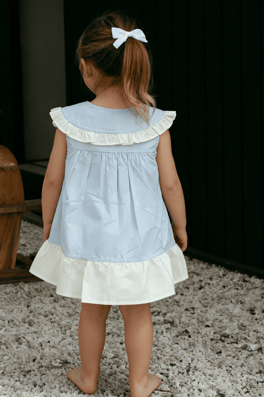 Child wearing a light blue dress with white ruffled details, standing on a textured surface.