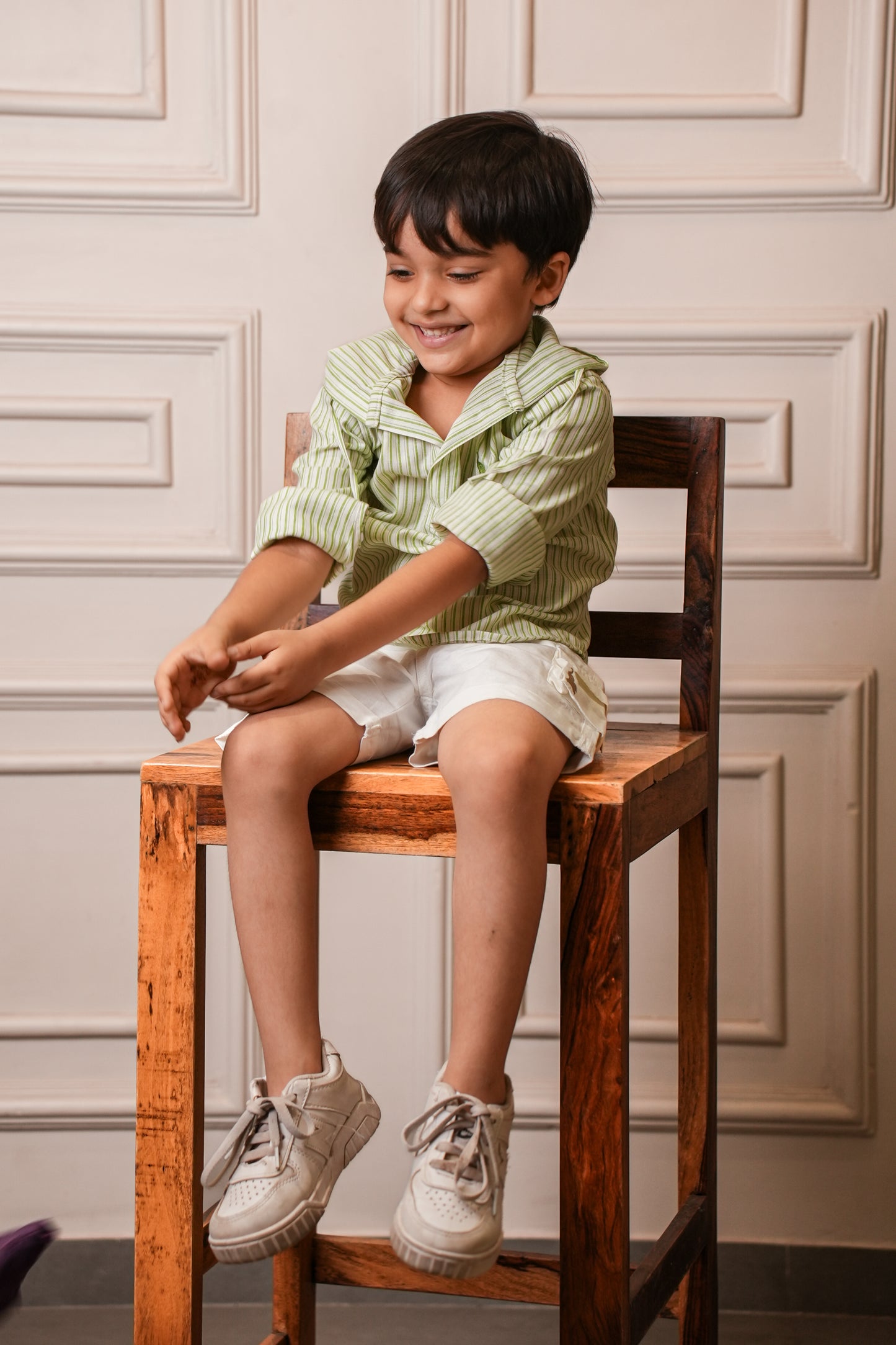 Child sitting on a wooden chair wearing a green and white striped shirt and white shorts.