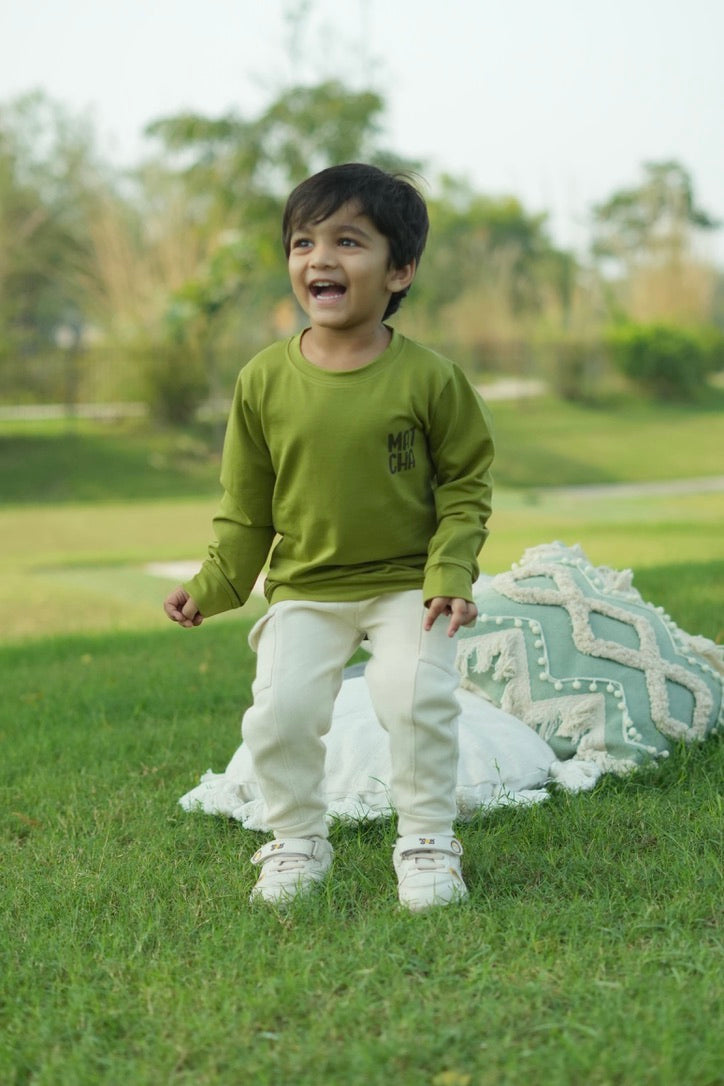 Child in a green shirt and white pants standing on grass with a blanket and pillow in the background
