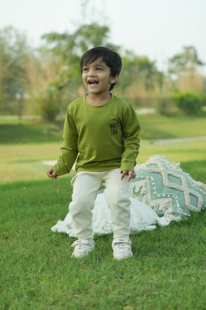 Child in a green shirt and white pants standing on grass with a blanket and pillow in the background