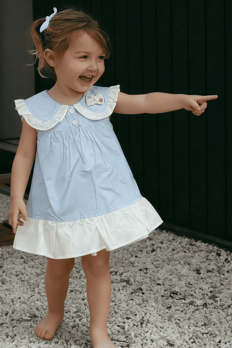 Young girl in a light blue dress with white ruffled collar and skirt, pointing forward.