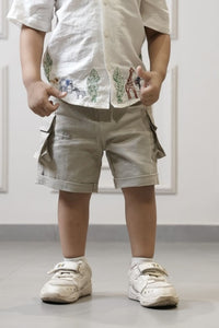 Child wearing a white shirt with embroidery and beige shorts, standing indoors.