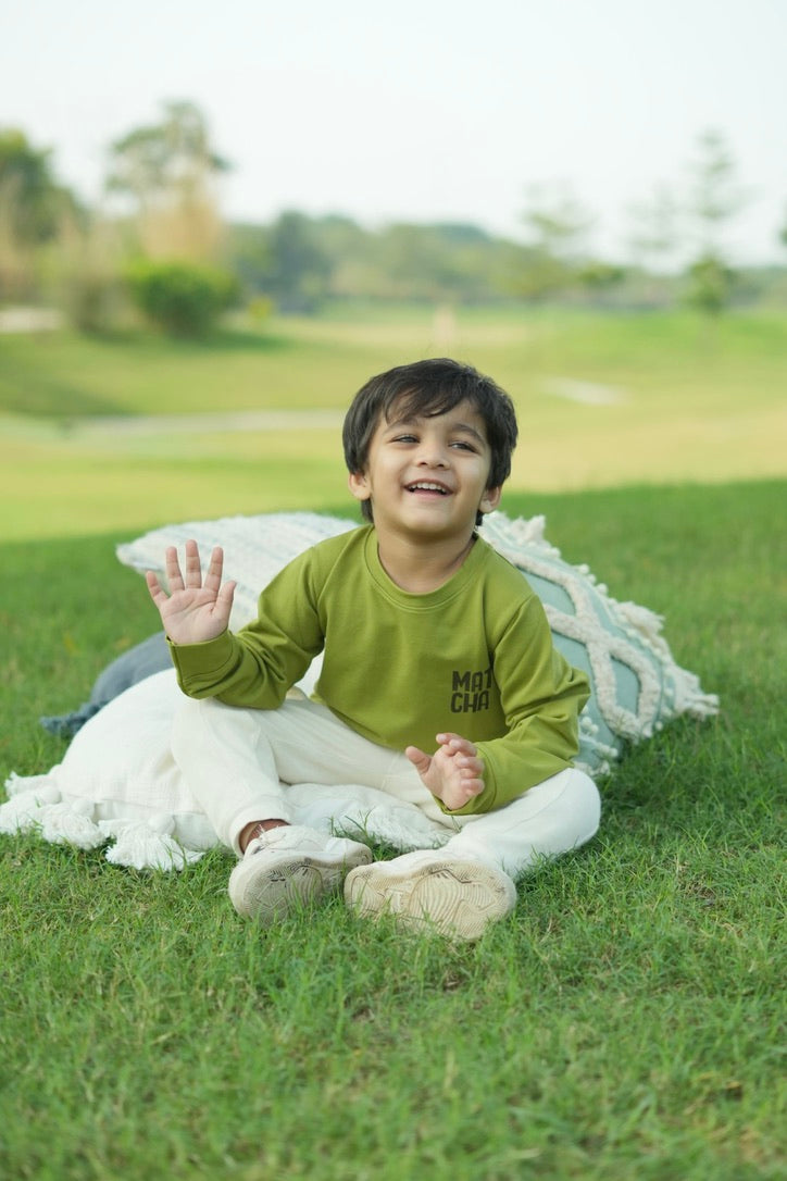Child sitting on grass wearing a green shirt with text, waving.