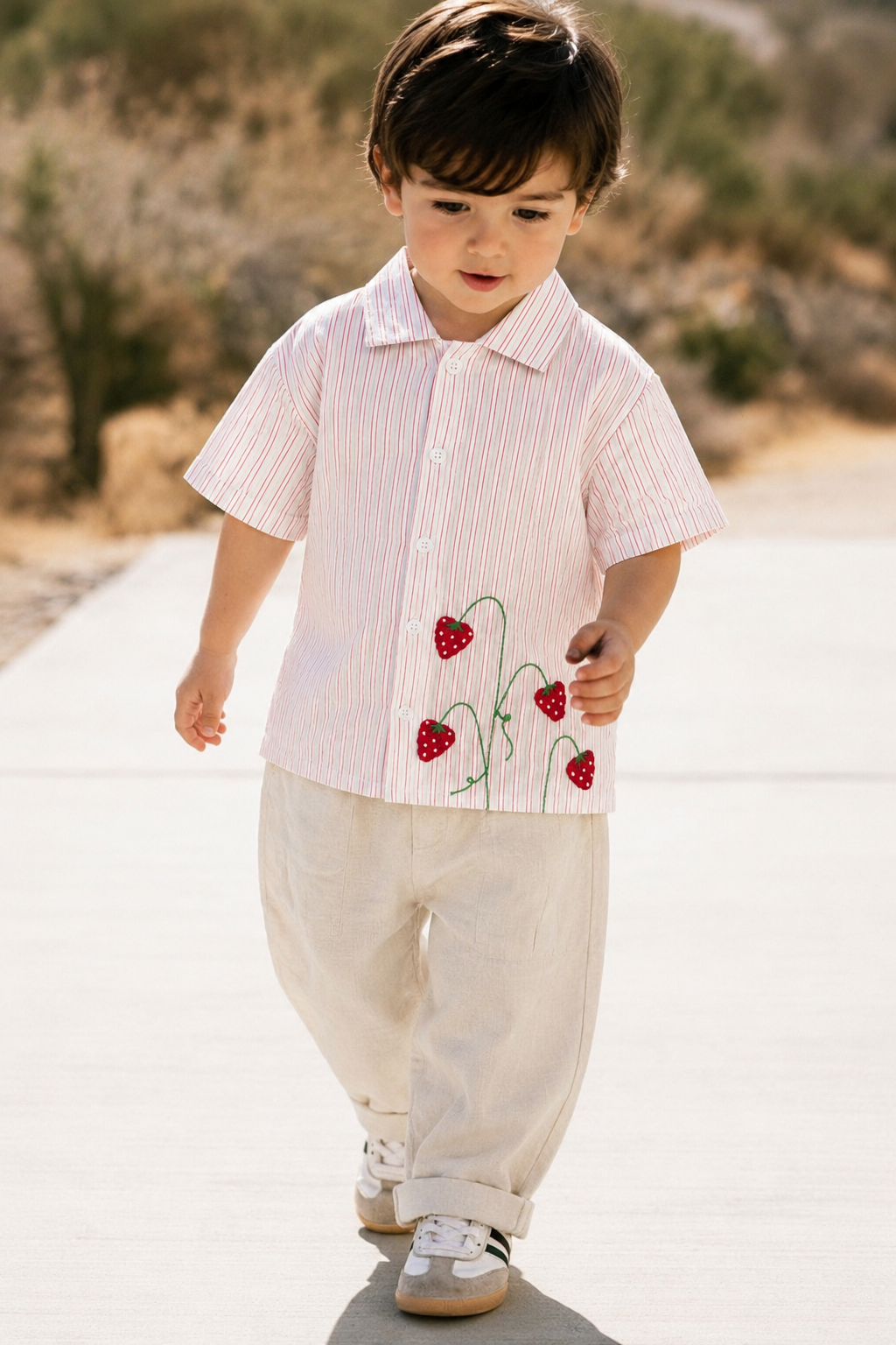 A toddler walking outdoors on a paved path wearing a short-sleeve collared shirt with thin vertical pink and white stripes and red strawberry embroidery, paired with cream pants and white sneakers.