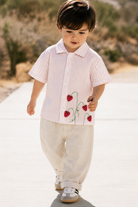 A toddler walking outdoors on a paved path wearing a short-sleeve collared shirt with thin vertical pink and white stripes and red strawberry embroidery, paired with cream pants and white sneakers.