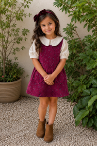 A young girl stands outdoors in a garden setting wearing a burgundy floral pinafore dress over a cream short-sleeve collared shirt, with a matching burgundy bow in her hair and brown ankle boots.