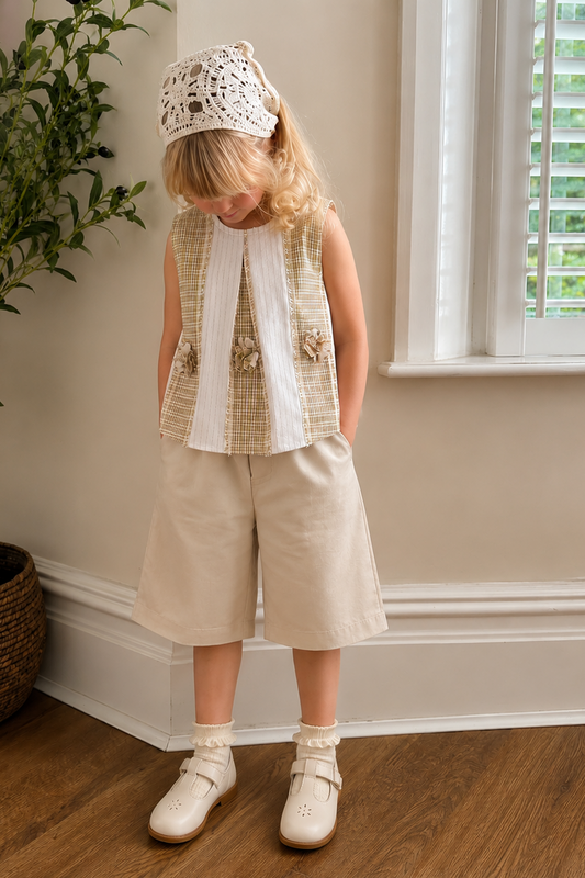 Young girl in a sleeveless checkered top and beige shorts standing in a room with a plant and window.