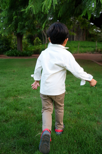 A young boy seen from behind running across a green lawn in a park, wearing a white long-sleeve button-up shirt, tan pants, and dark sneakers with bright red accents.