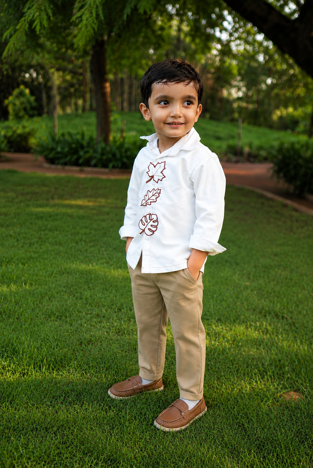 A young boy standing on a green lawn wearing a white long-sleeve button-up shirt with brown embroidered leaf motifs, khaki pants, and brown loafers.