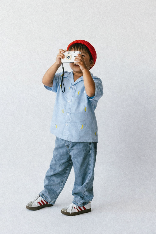 Child holding a camera, wearing a blue shirt with yellow buttons and jeans, on a plain background