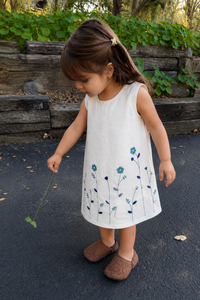 A young girl wearing a sleeveless white dress with blue floral embroidery, standing outdoors on a paved surface in front of a rustic wooden wall and green foliage.