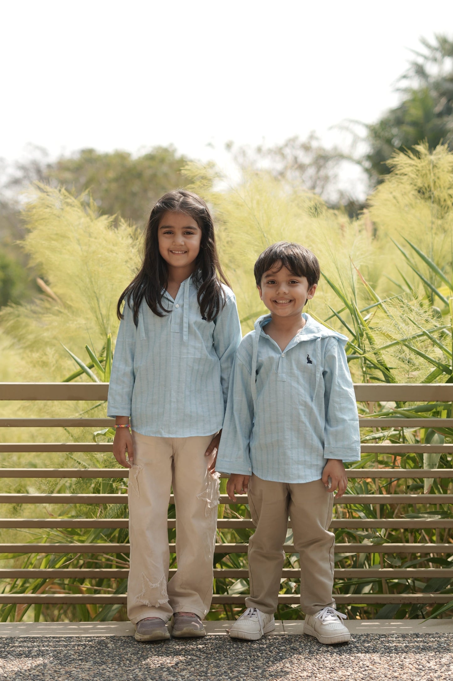 Two kids standing outdoors with a wooden fence and greenery in the background