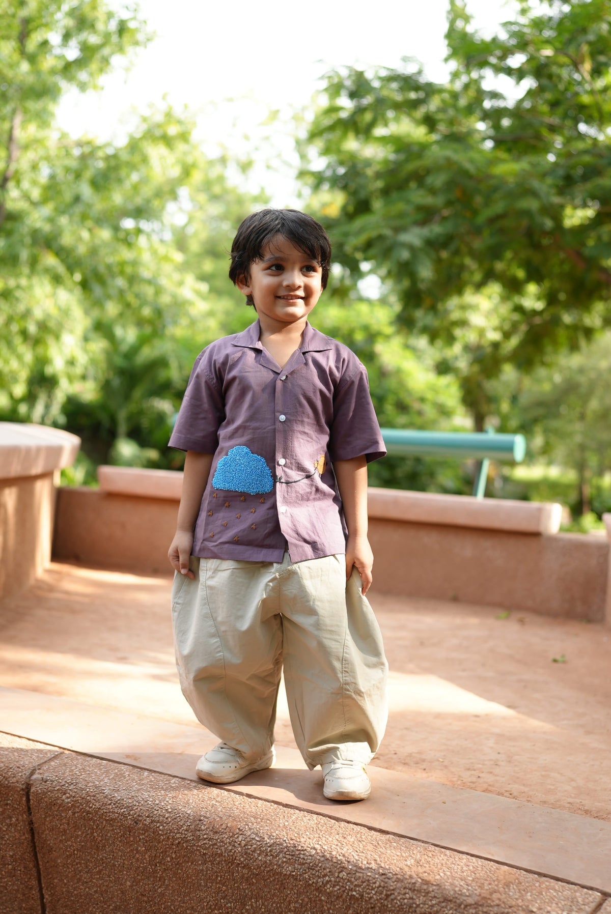 A young boy wearing a purple short-sleeve polo shirt with a blue heart graphic, beige trousers, and white sneakers, standing outdoors on a stone walkway with green trees in the background.