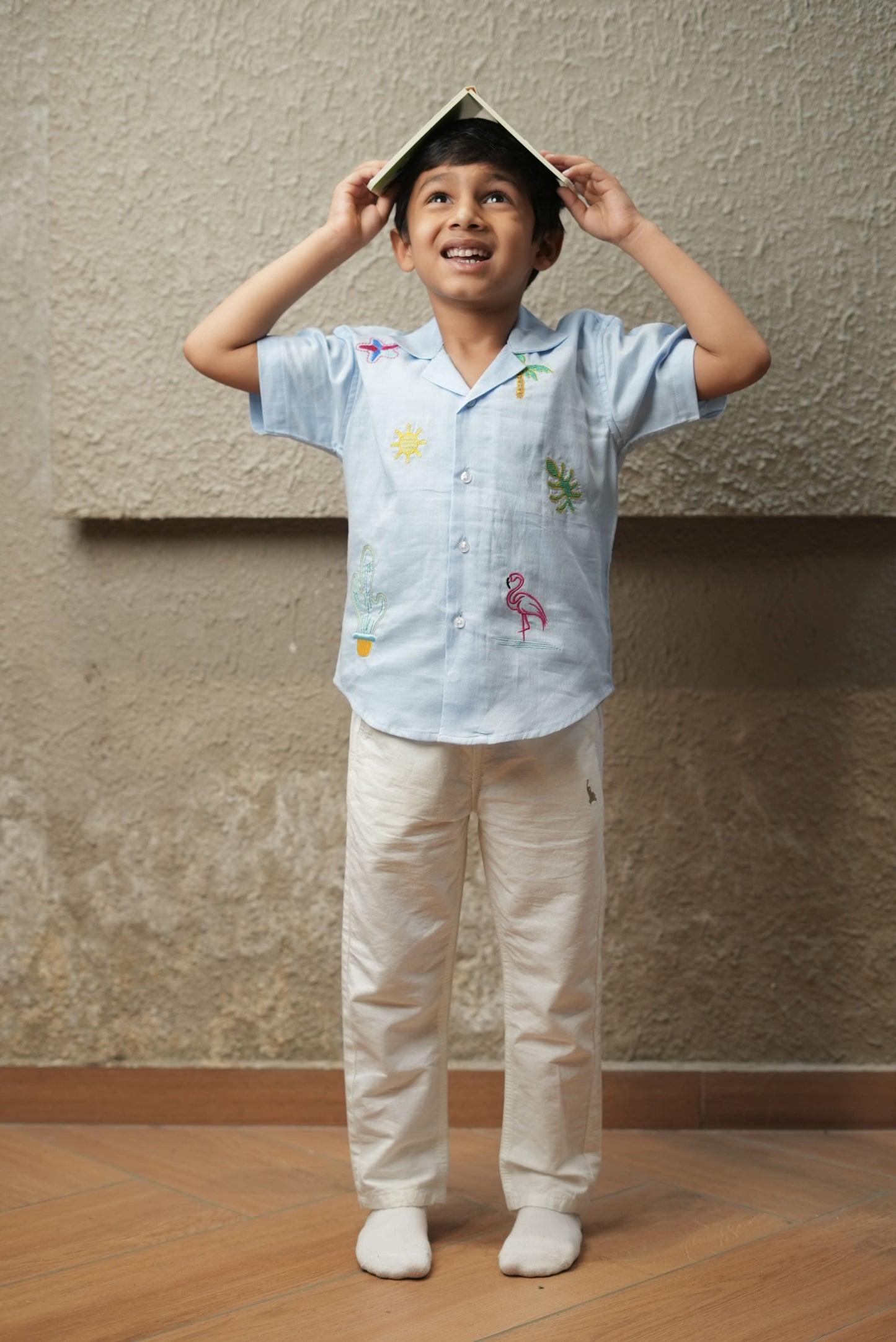 Child wearing a light blue shirt with colorful designs and beige pants, standing against a plain wall.