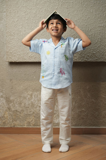 Child wearing a light blue shirt with colorful designs and beige pants, standing against a plain wall.