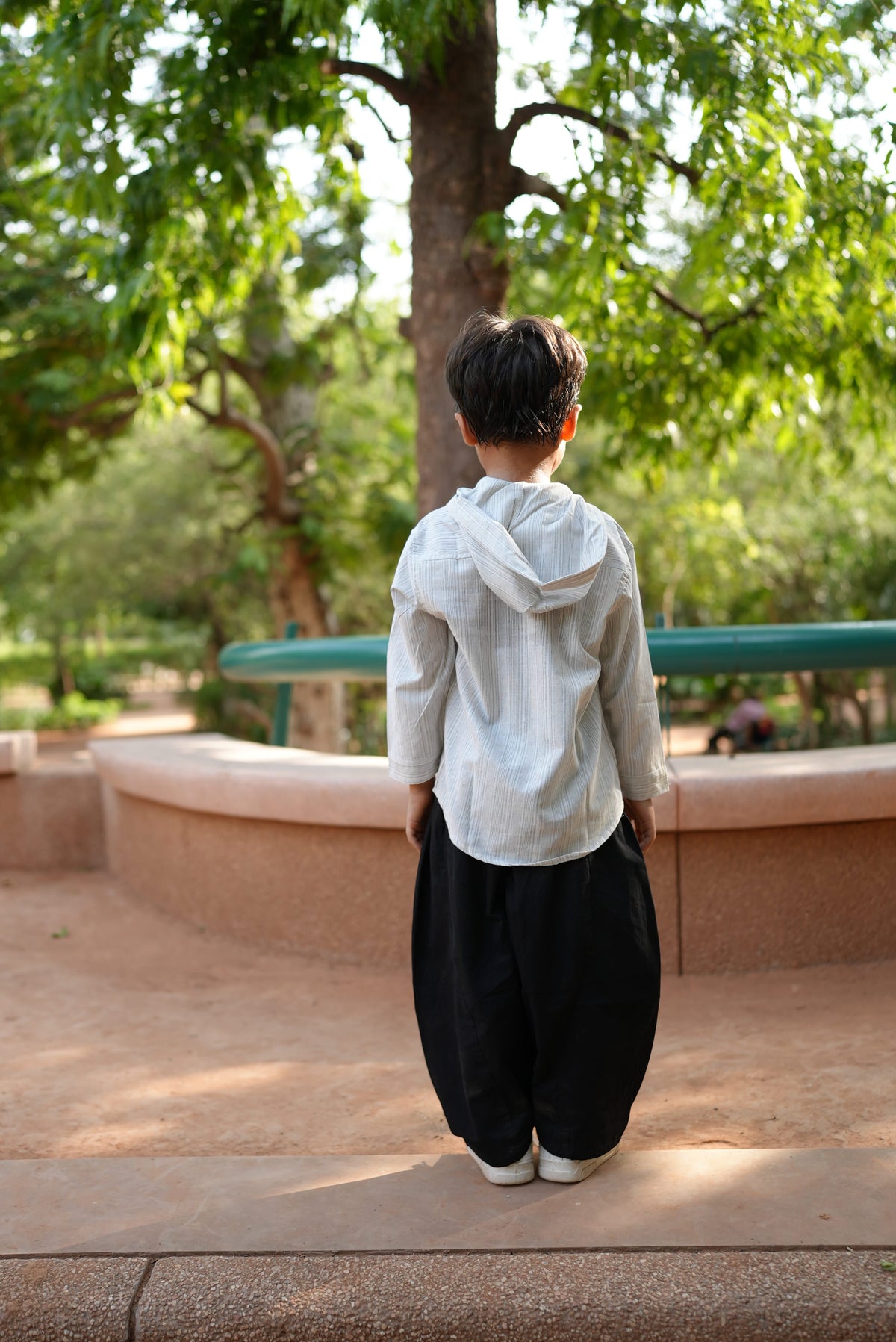 A person wearing a white long-sleeve shirt and black pants stands outdoors on a paved walkway with trees and greenery in the background.