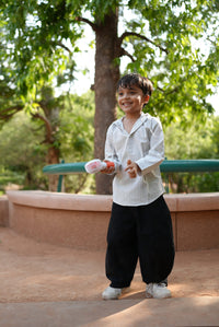 A young boy wearing a white long-sleeve shirt and black pants stands outdoors in a park-like setting with trees and a tan stone structure behind him, holding a small white bottle.