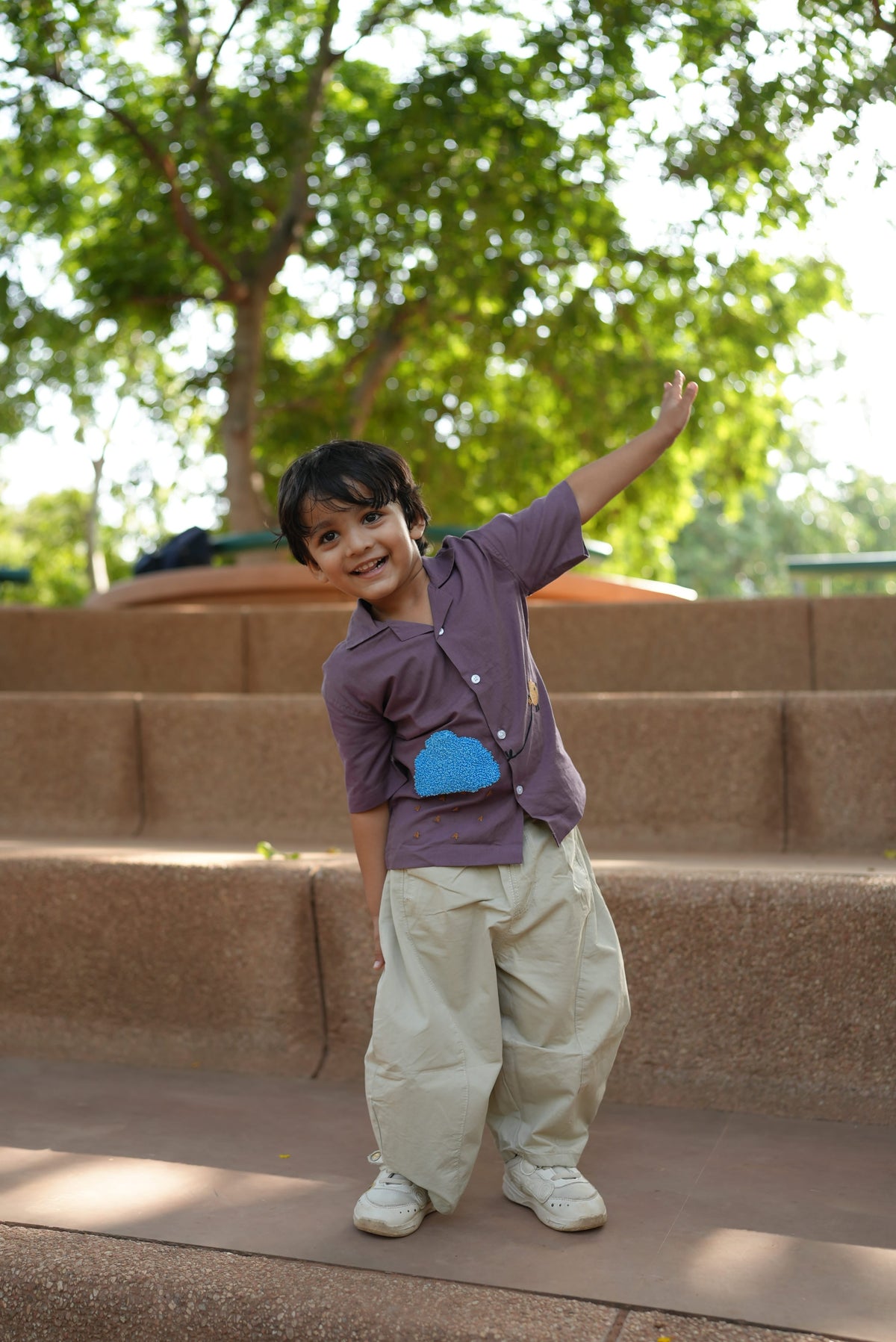 A young boy smiling and posing outdoors on stone steps, wearing a purple short-sleeve button-up shirt with a blue heart detail, beige cargo pants, and white sneakers.