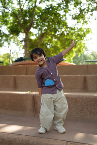A young boy smiling and posing outdoors on stone steps, wearing a purple short-sleeve button-up shirt with a blue heart detail, beige cargo pants, and white sneakers.