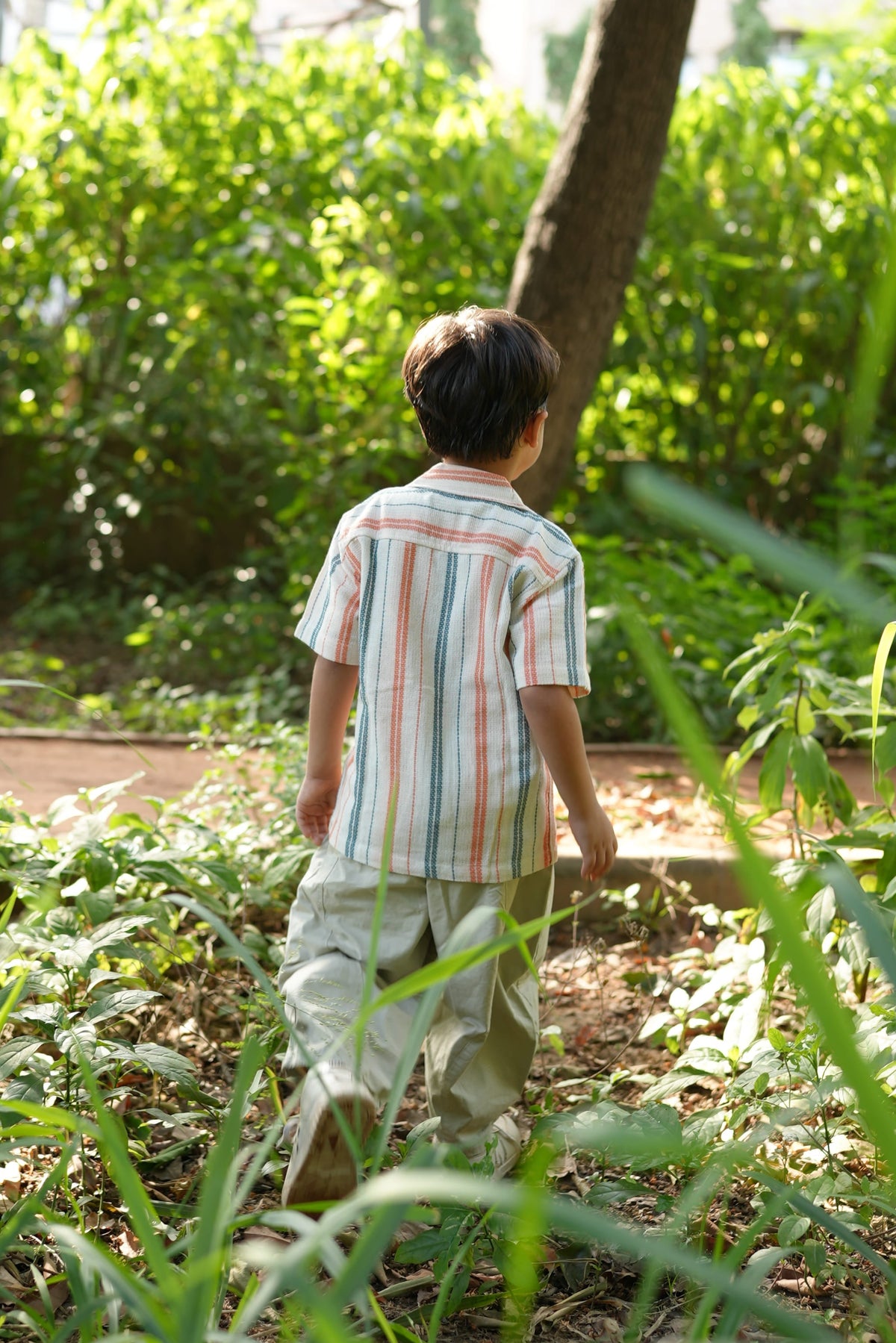 A child wearing a striped short-sleeve shirt and light-colored pants bends down in a lush green garden.