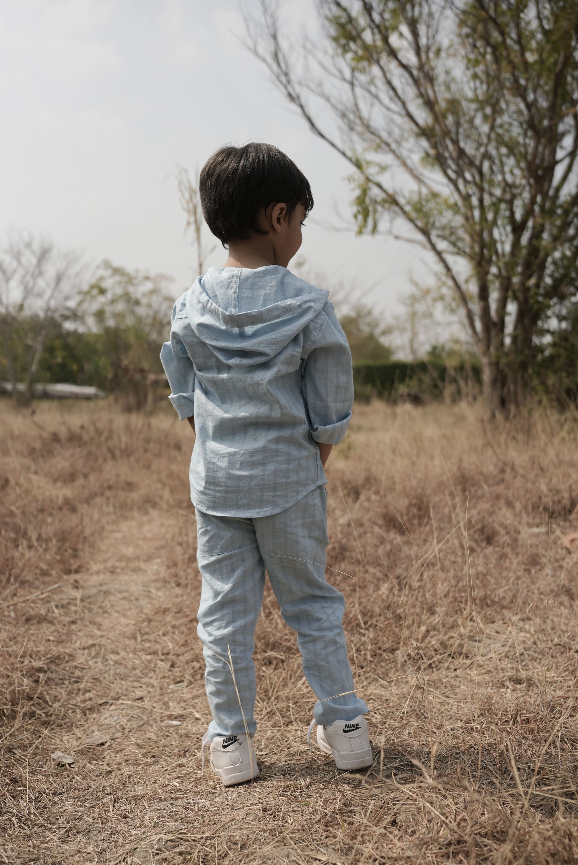 Child in a light blue outfit standing in a dry field with trees in the background