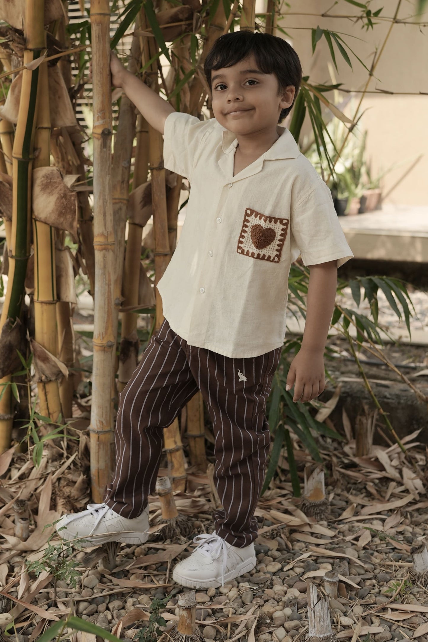 Child standing among bamboo plants wearing a white shirt with a design and brown pants.