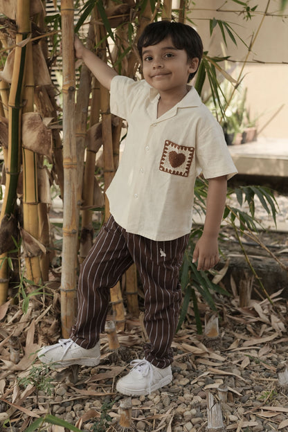 Child standing among bamboo plants wearing a white shirt with a design and brown pants.