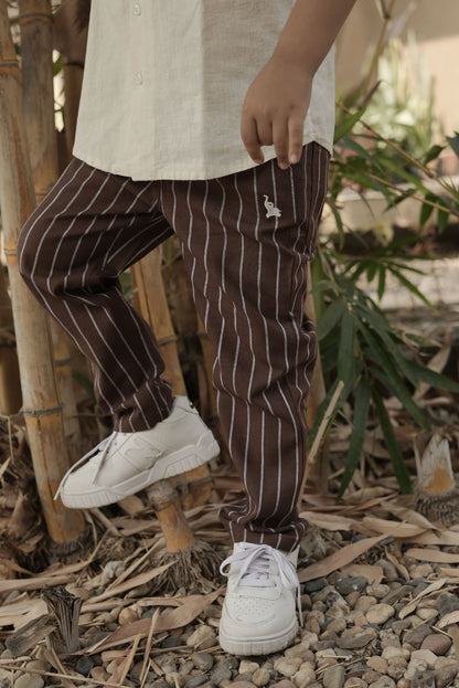 Person wearing brown pinstripe pants and white sneakers standing on a natural ground with plants in the background.
