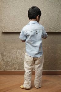 Child wearing a light blue shirt and beige pants standing against a plain wall.