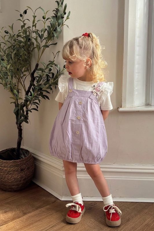 Child wearing a purple dress with white buttons, standing indoors near a plant and window.