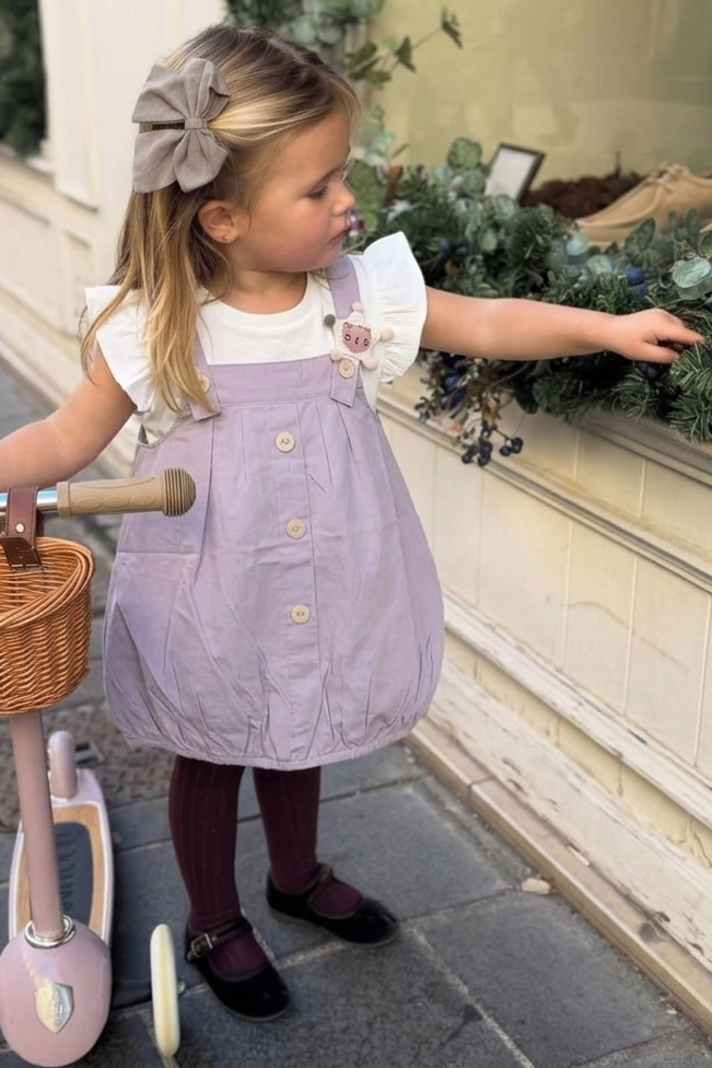 Child in a purple dress standing next to a pink scooter with a basket on a sidewalk.