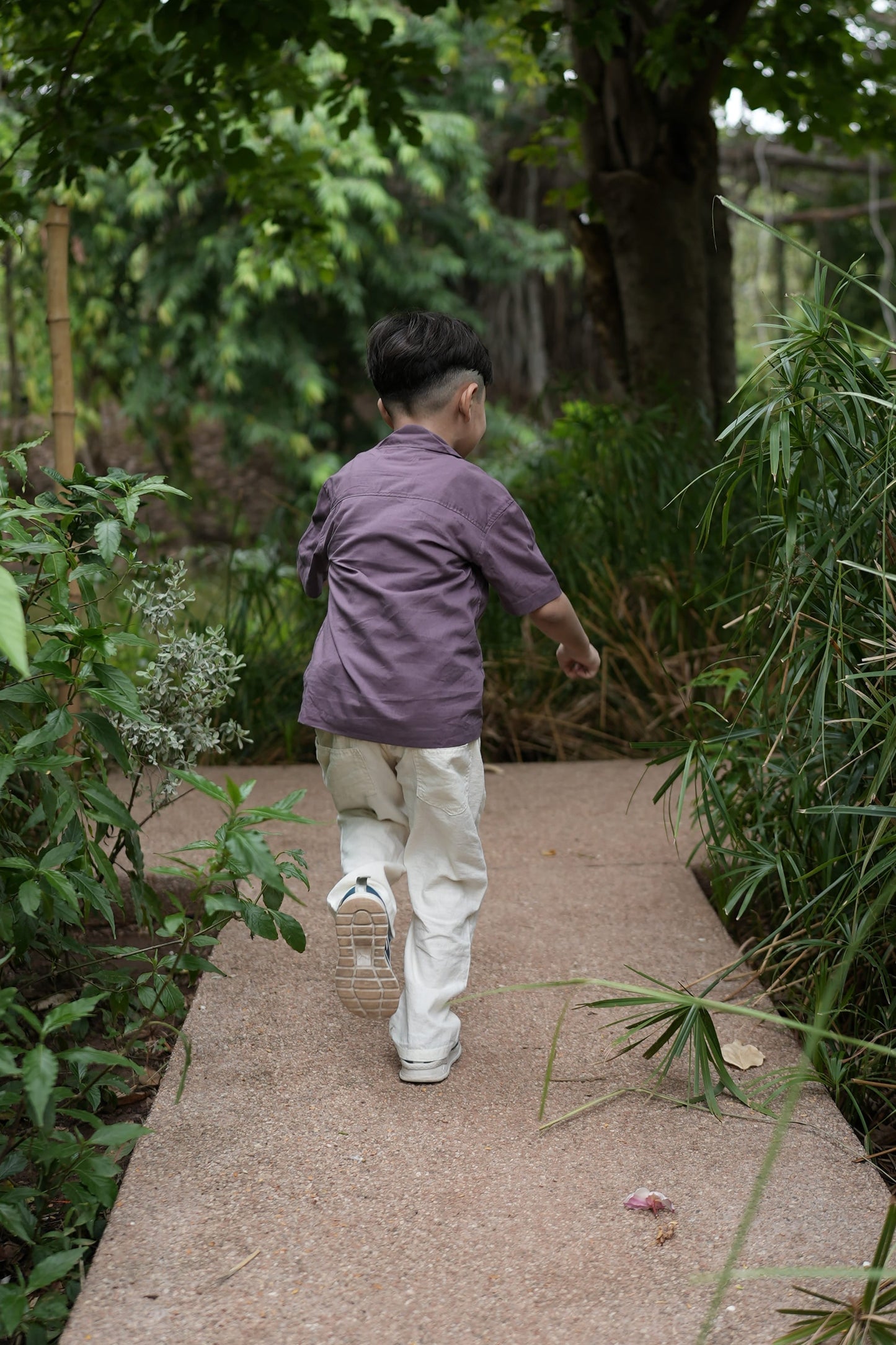 A kid wearing a purple long-sleeve top and off-white pants walks along a concrete path surrounded by green plants and trees.