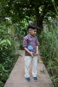 A young boy wearing a purple short-sleeve button-up shirt with a blue heart on the chest, off-white trousers, and blue sneakers, standing outdoors on a stone path surrounded by green plants.