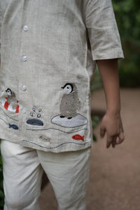A close-up of a kid wearing a light beige linen kurta with embroidered penguins, fish, and a polar bear on the chest, paired with matching beige trousers.