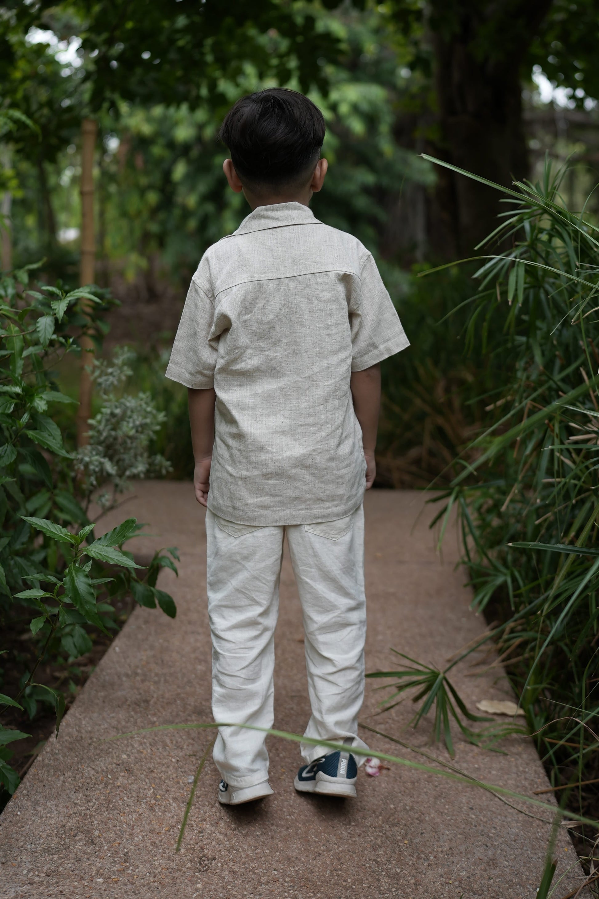 A child viewed from behind wearing a light beige linen short-sleeve shirt and matching linen trousers, standing on a stone path surrounded by green plants.
