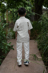 A child viewed from behind wearing a light beige linen short-sleeve shirt and matching linen trousers, standing on a stone path surrounded by green plants.