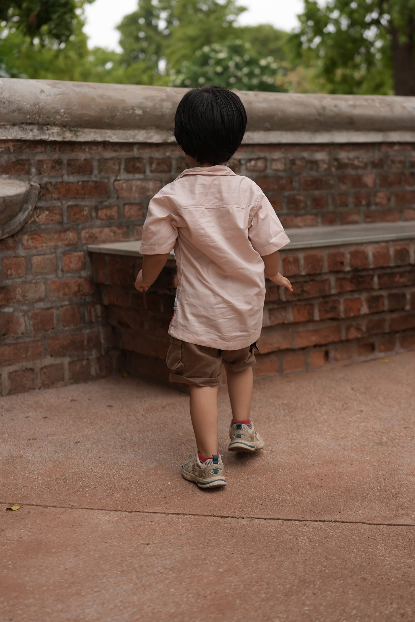 A child wearing a light pink t-shirt, brown shorts, and sneakers stands on a brick and concrete structure outdoors with trees in the background.