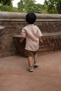 A child wearing a light pink t-shirt, brown shorts, and sneakers stands on a brick and concrete structure outdoors with trees in the background.